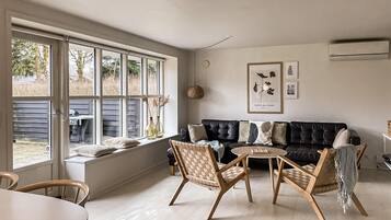 Un salon moderne avec un canapé en cuir noir, des chaises en bois, une petite table ronde et de grandes fenêtres donnant sur un jardin.