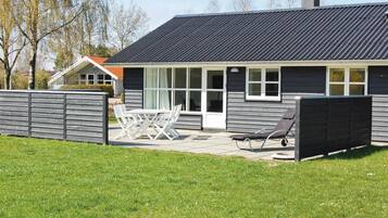 Une maison moderne avec un toit foncé, de grandes fenêtres et une terrasse aménagée avec du mobilier d’extérieur.