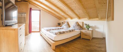 Un dormitorio con techo de madera, una cama con ropa de cama estampada, un televisor sobre un tocador de madera y una mesita de noche con una planta.