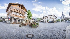 A cobblestone square with a wooden building featuring balconies and a church with a dome in the background.
