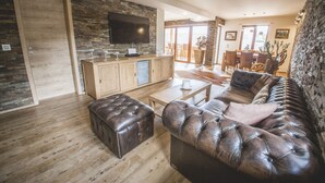 A modern living room with a wooden floor, a large leather sofa, a television mounted on a stone wall, and a dining area with wooden furniture.