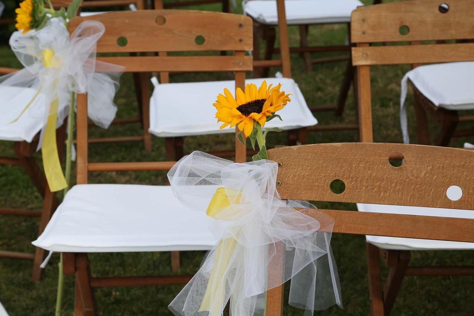 Reihenweise Holzsessel mit weißen Tischdecken und gelben Blumen, die mit weißen Bändern verbunden sind.
