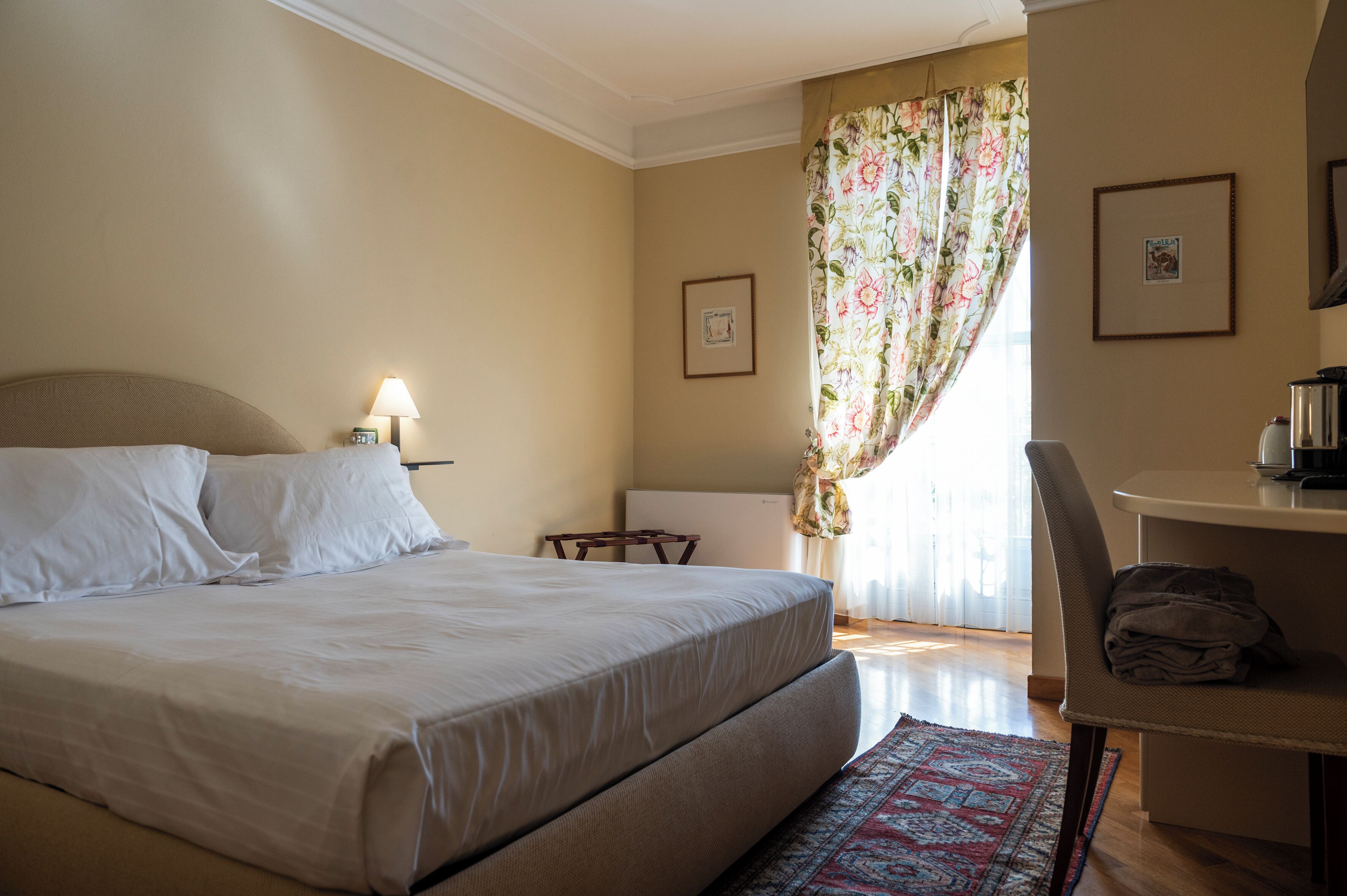 Quarto de hotel com cama, escrivaninha, cadeira, luminária, radiador e janela com cortinas.