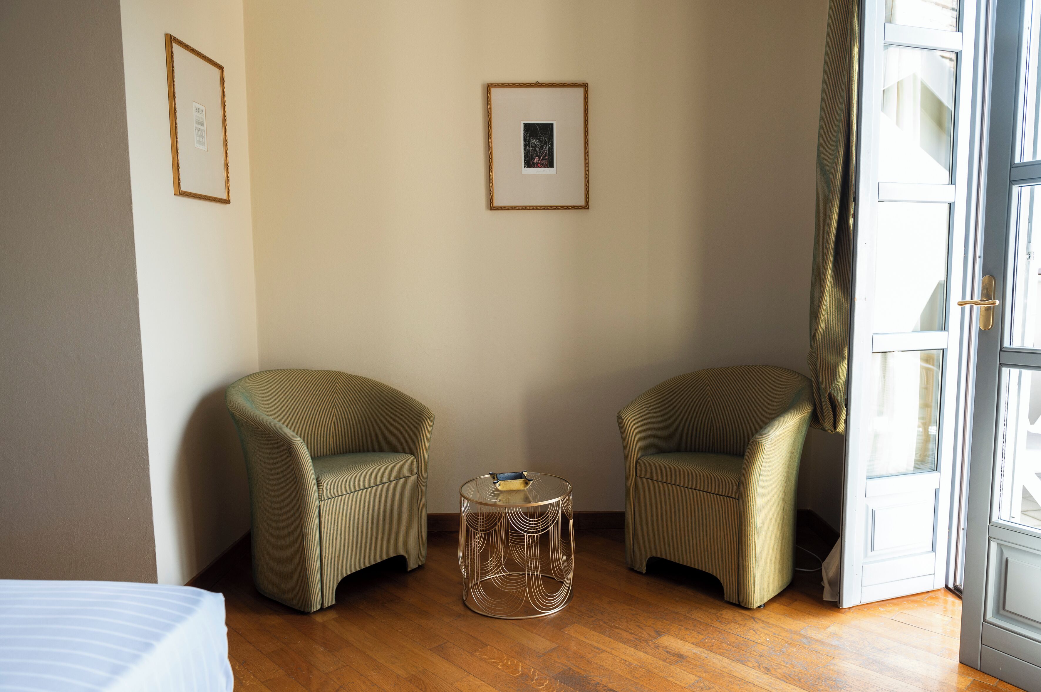 Une pièce comprenant deux fauteuils verts à revêtement en tissu, une table basse ronde en verre et un lit recouvert d’une courtepointe à rayures.