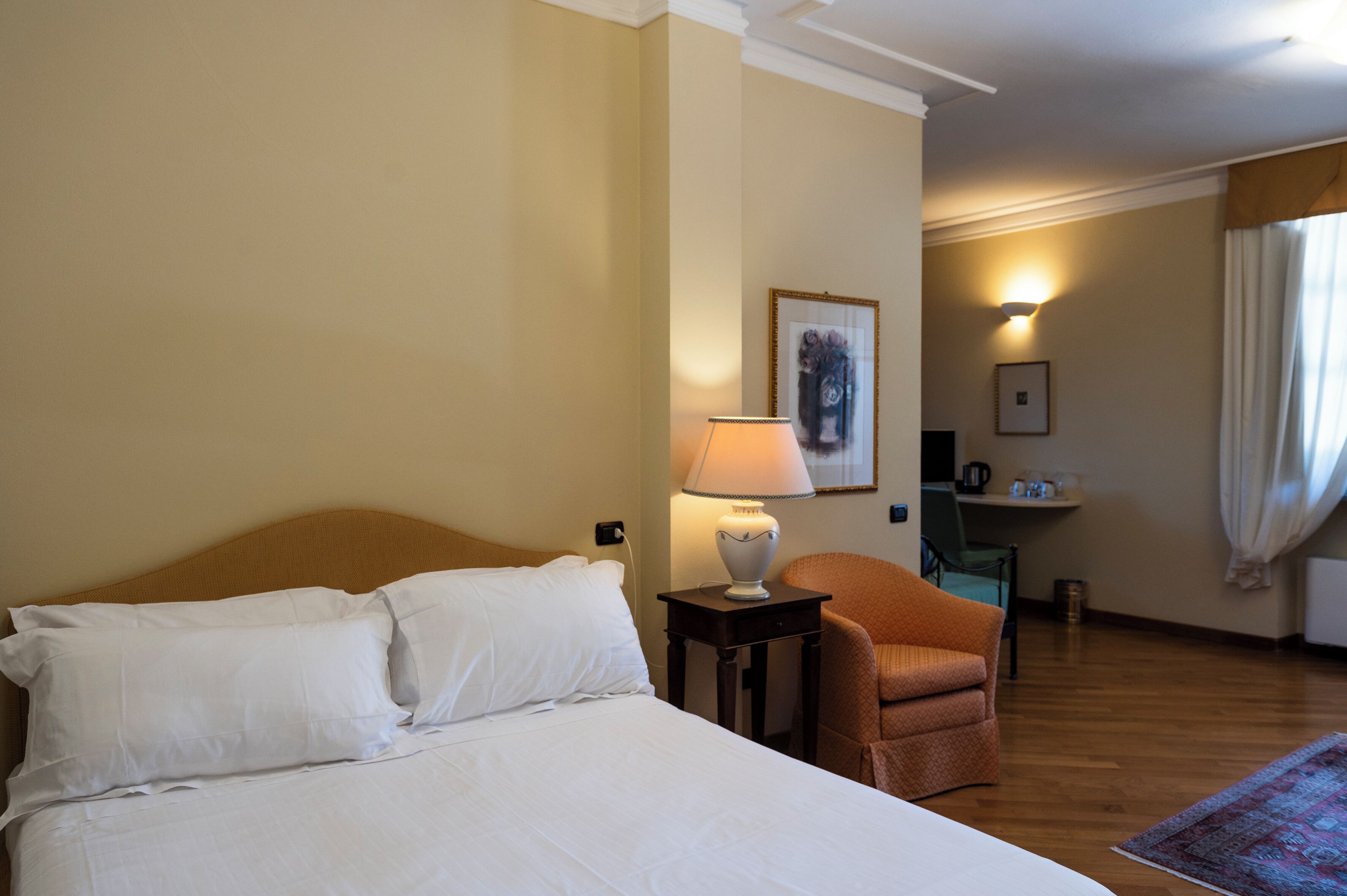 Ein Hotelzimmer mit Bett, Lampe, Sessel, Schreibtisch und einem gerahmten Bild an der Wand.