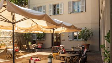 Una terrazza esterna con tavoli e sedie, pavimento in pietra e un edificio con diverse finestre e persiane.