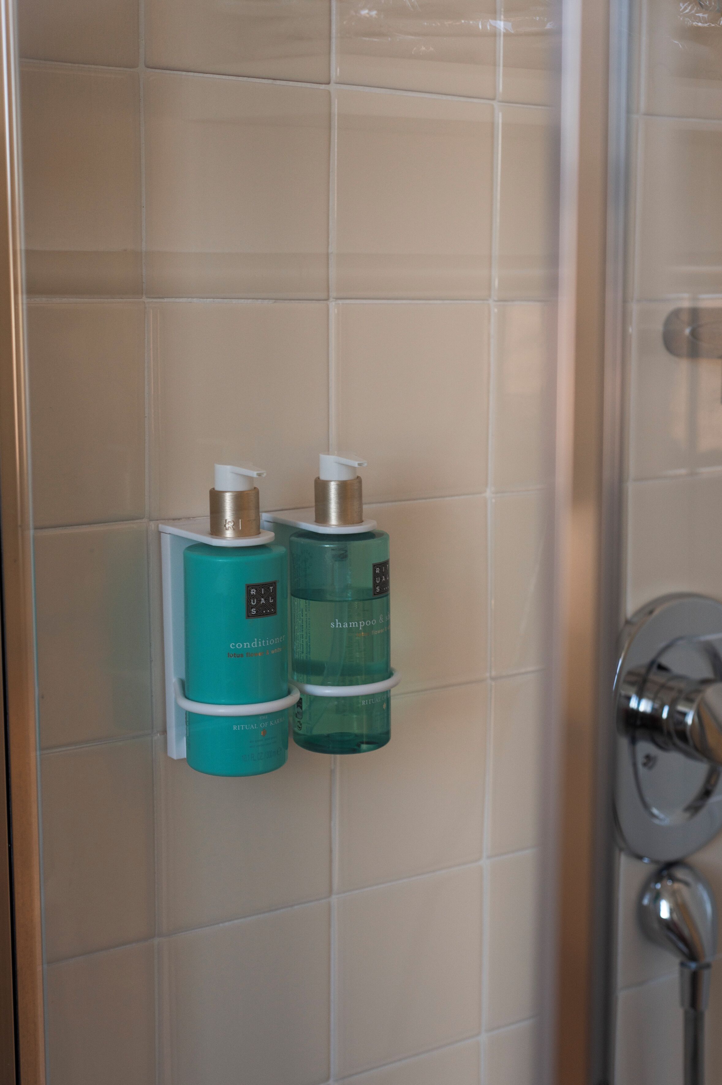 A bathroom with beige tiles, a shower, and two teal bottles labeled 'Rituals' on a wall-mounted holder.