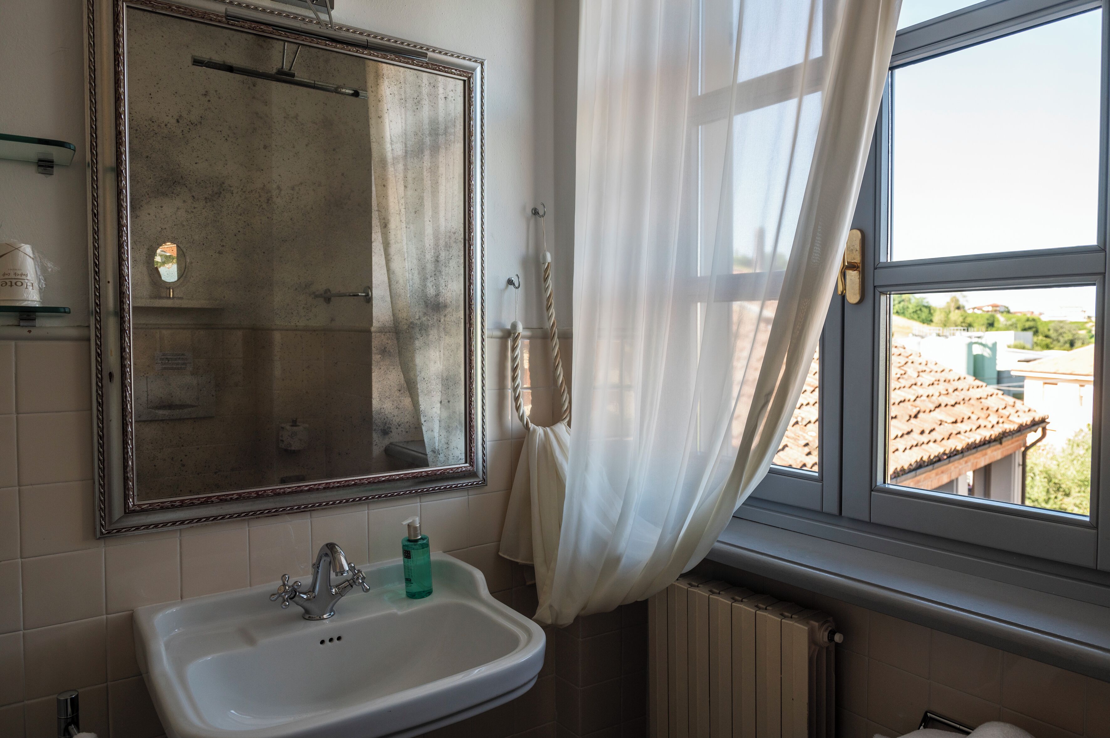 Ein Badezimmer mit einem großen Spiegel, einem Waschbecken mit Armatur, einem Fenster mit Blick auf Gebäude und einem Heizkörper.