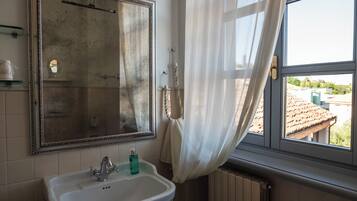 Ein Badezimmer mit einem großen Spiegel, einem Waschbecken mit Armatur, einem Fenster mit Blick auf Gebäude und einem Heizkörper.