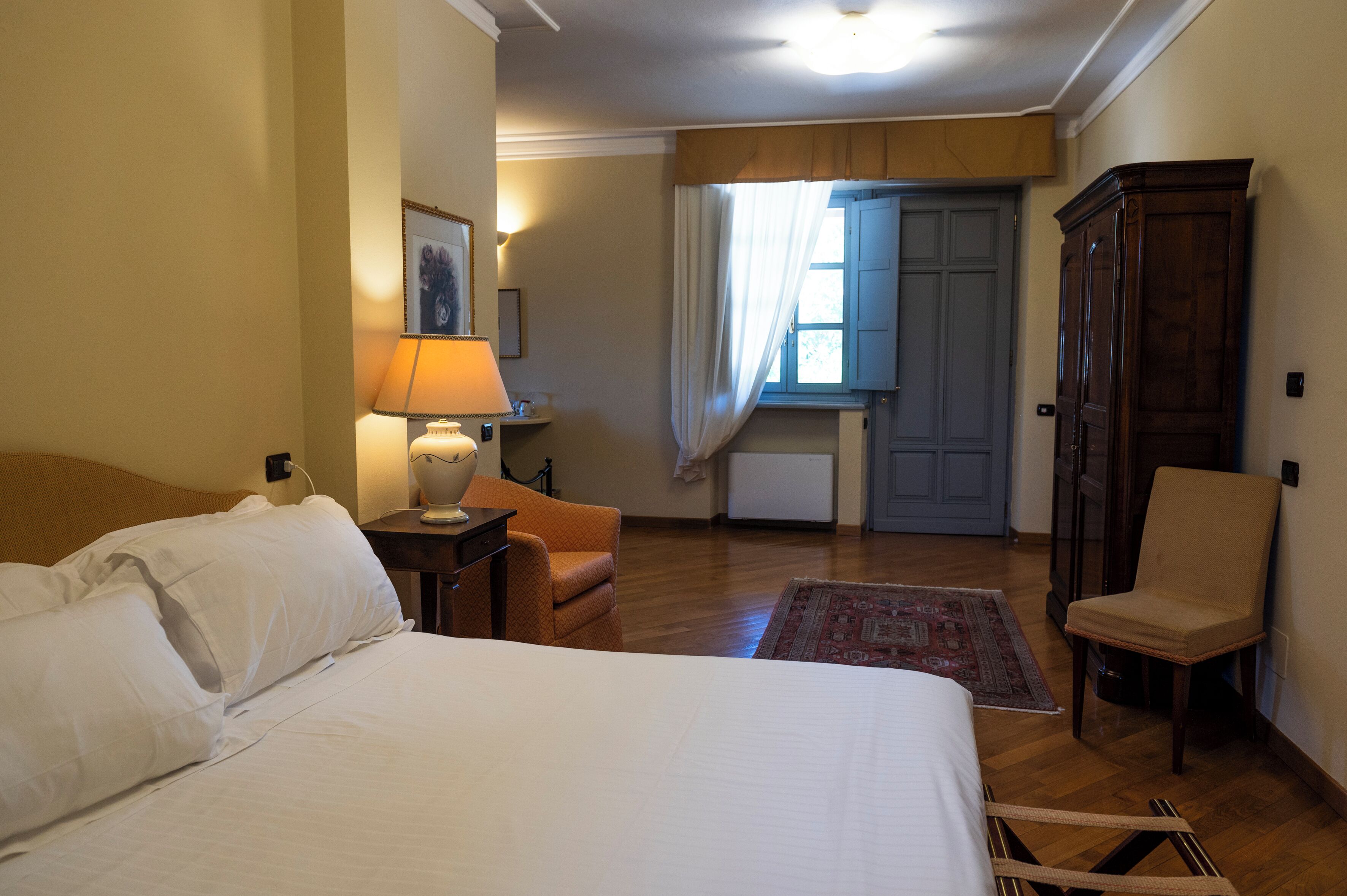 A hotel room with a bed, a chair, a desk, a lamp, a wardrobe, and a window with curtains.