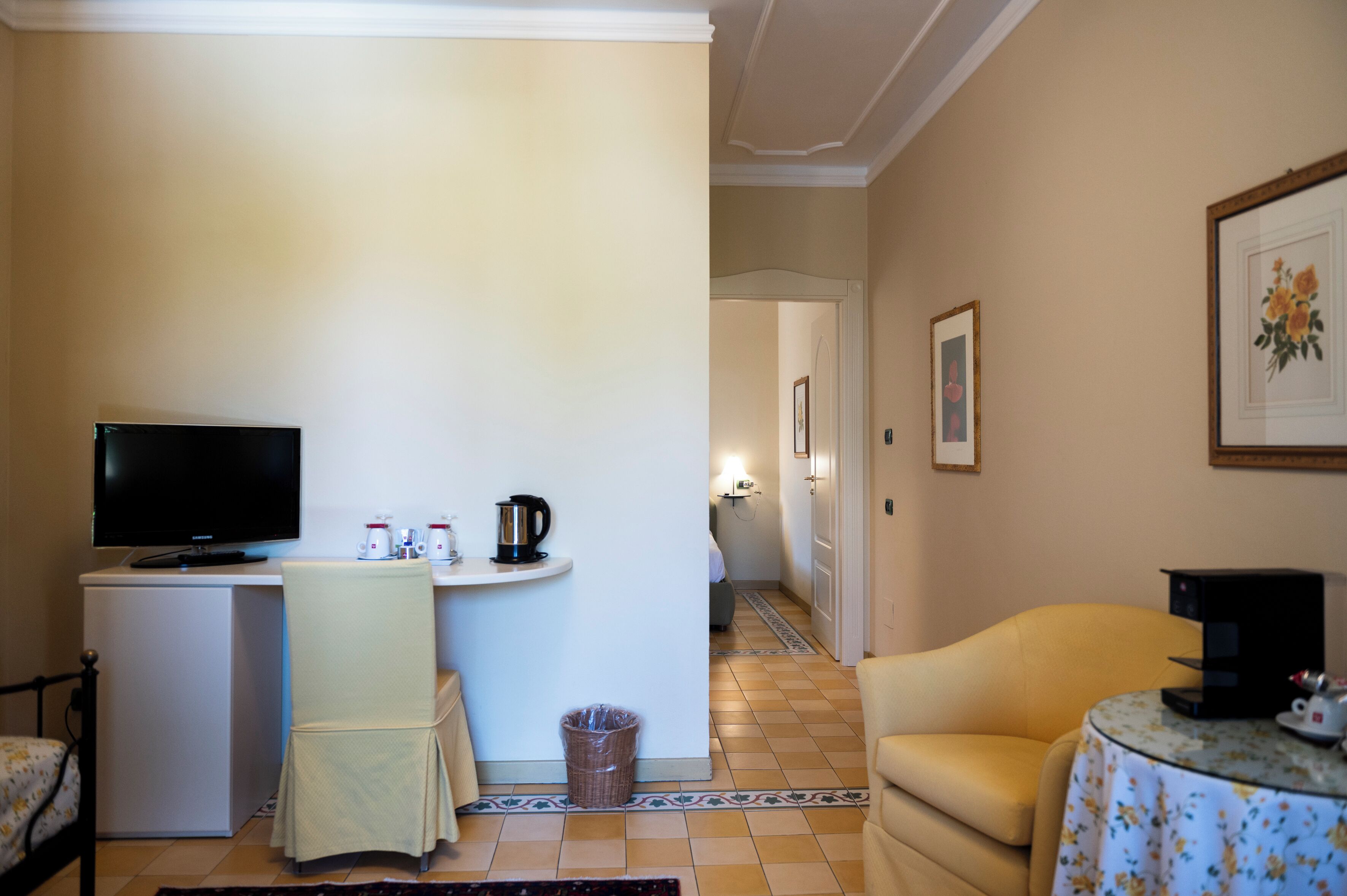 A hotel room with a TV, a small table with a kettle, a chair, a sofa, and a trash bin.