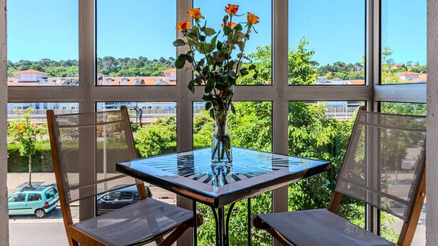 A table with a vase of flowers, two chairs, and a view of buildings and greenery through large windows. - Convenient Arcachon T2 Apt - Balcony, Elevator, Secure Garage Near Shops & Train Station (Arcachon)