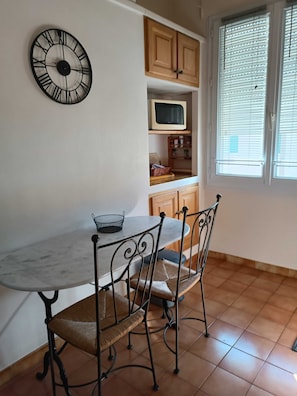 A dining area with a marble table and metal chairs, a wall clock, wooden cabinets, a microwave, and a window with blinds. - Homerez - Apartment in Arles (Arles)