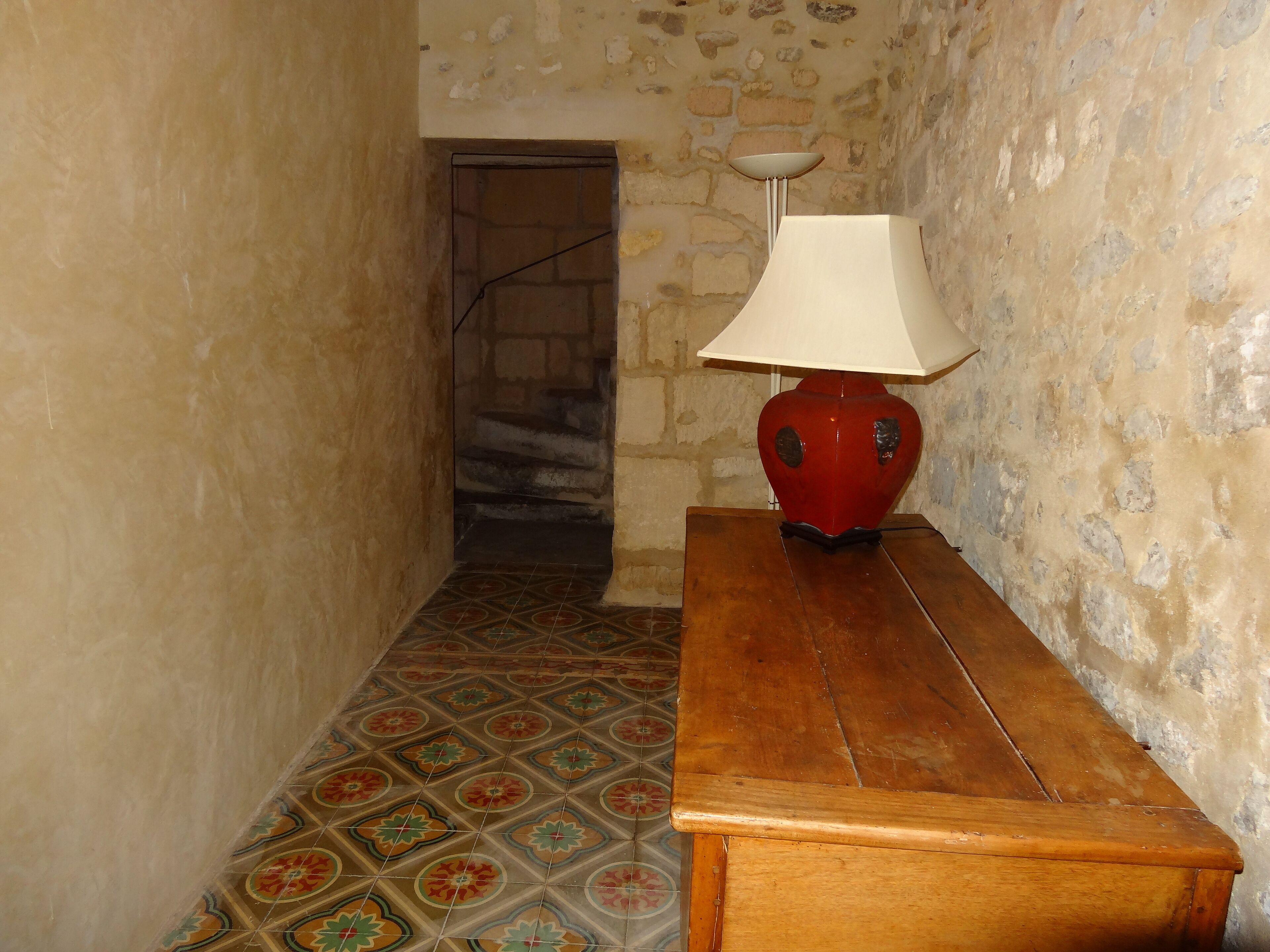 Ein schmaler Gang mit einer hölzernen Bank und einer roten Lampe mit weißem Schirm. Die Wände bestehen aus freiliegendem Mauerwerk, und der Boden ist mit gemusterten Fliesen ausgelegt.