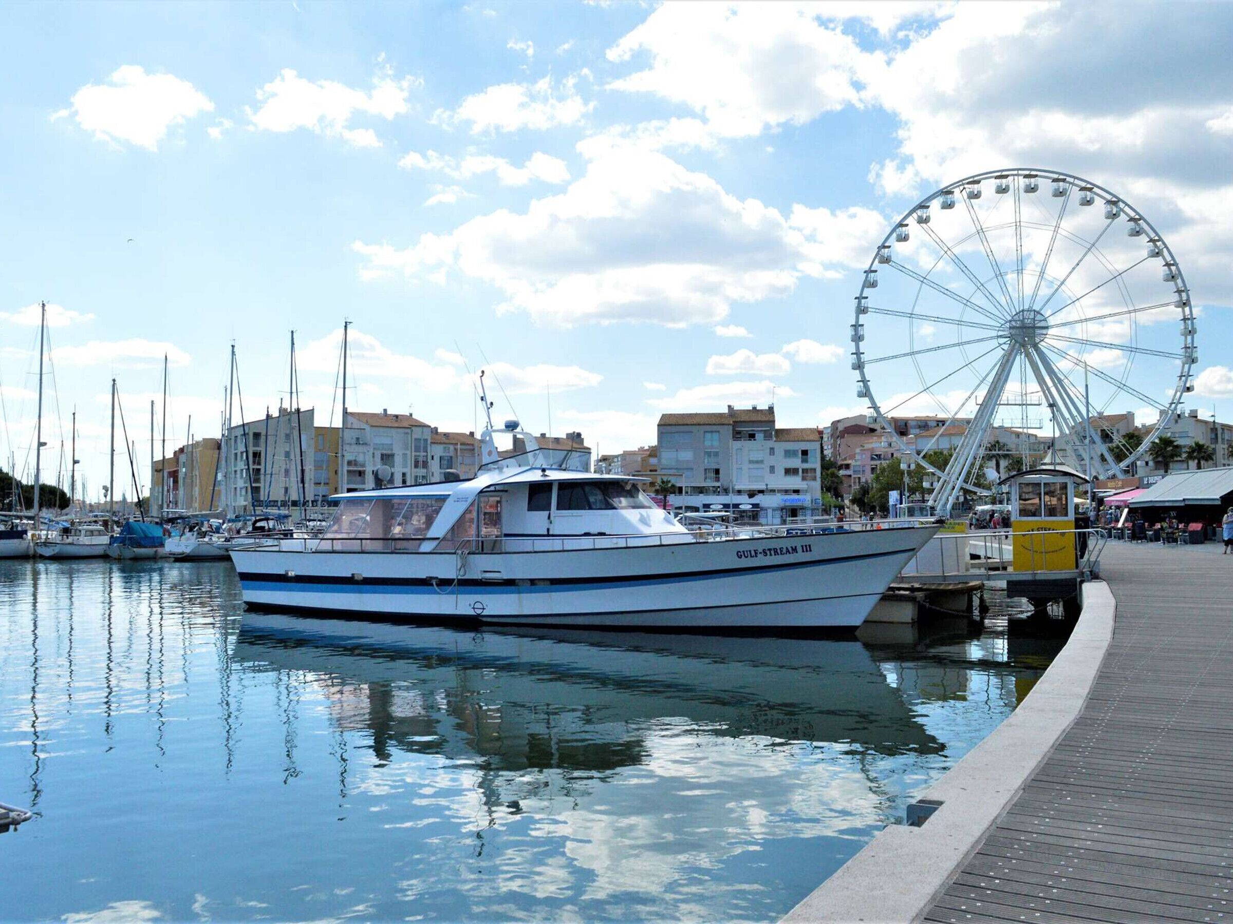 A marina with boats docked, a Ferris wheel in the background, and buildings along the waterfront.