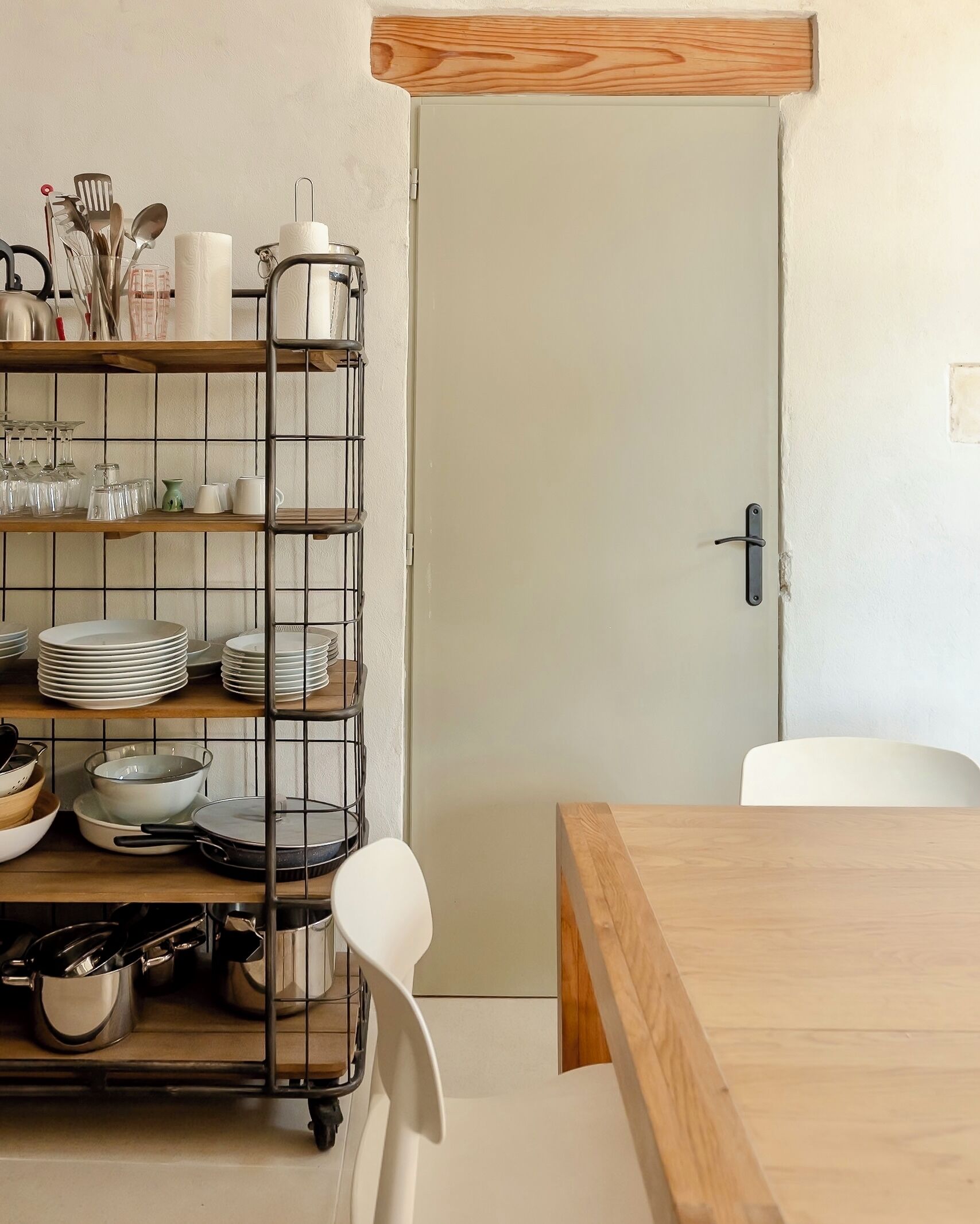 A kitchen with a wooden table and chairs, a metal rack with dishes, and a white door.
