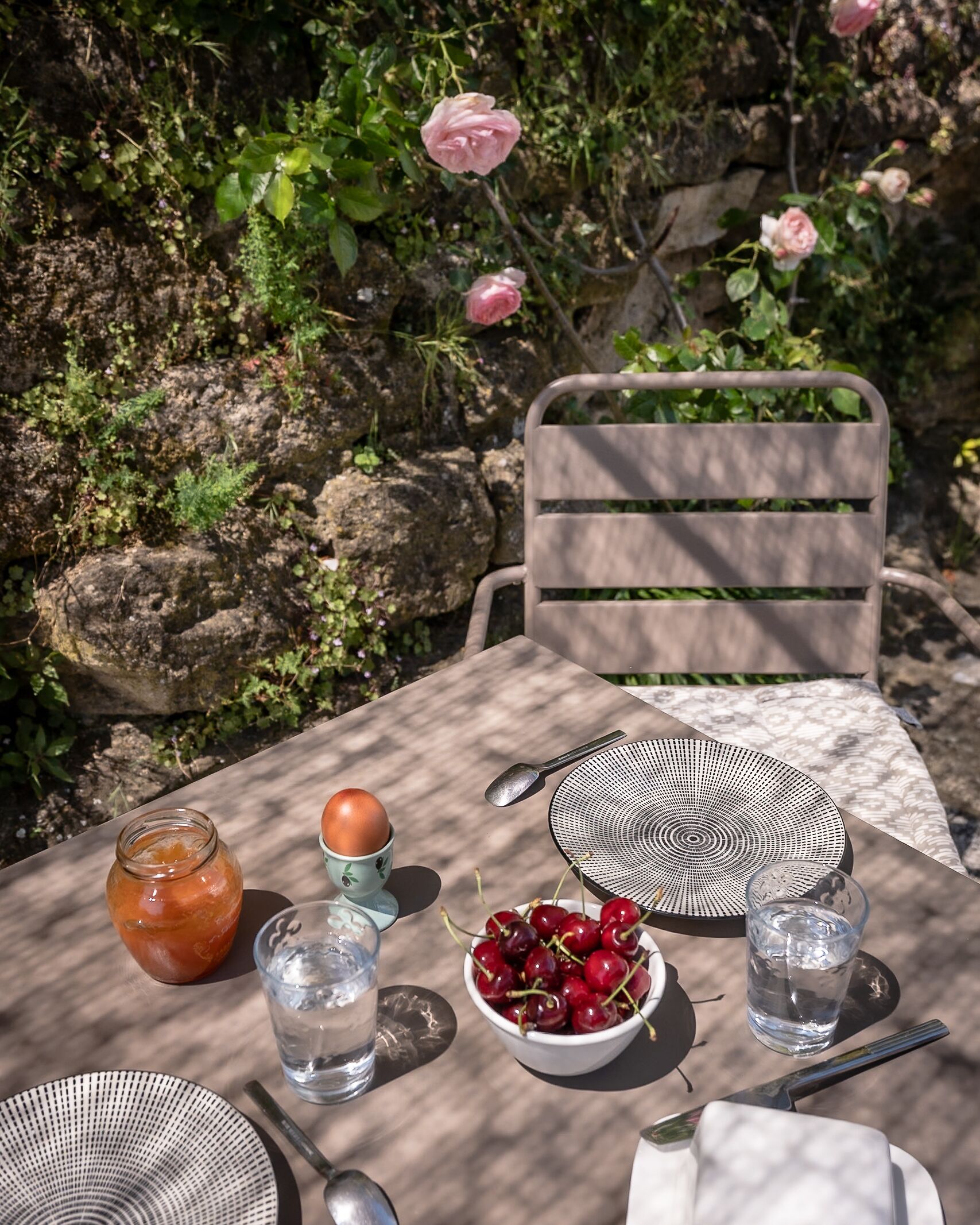 Mesa al aire libre con una silla rústica, un tazón de cerezas, un vaso de agua, una cuchara de metal, una taza de cerámica y un plato estampado.