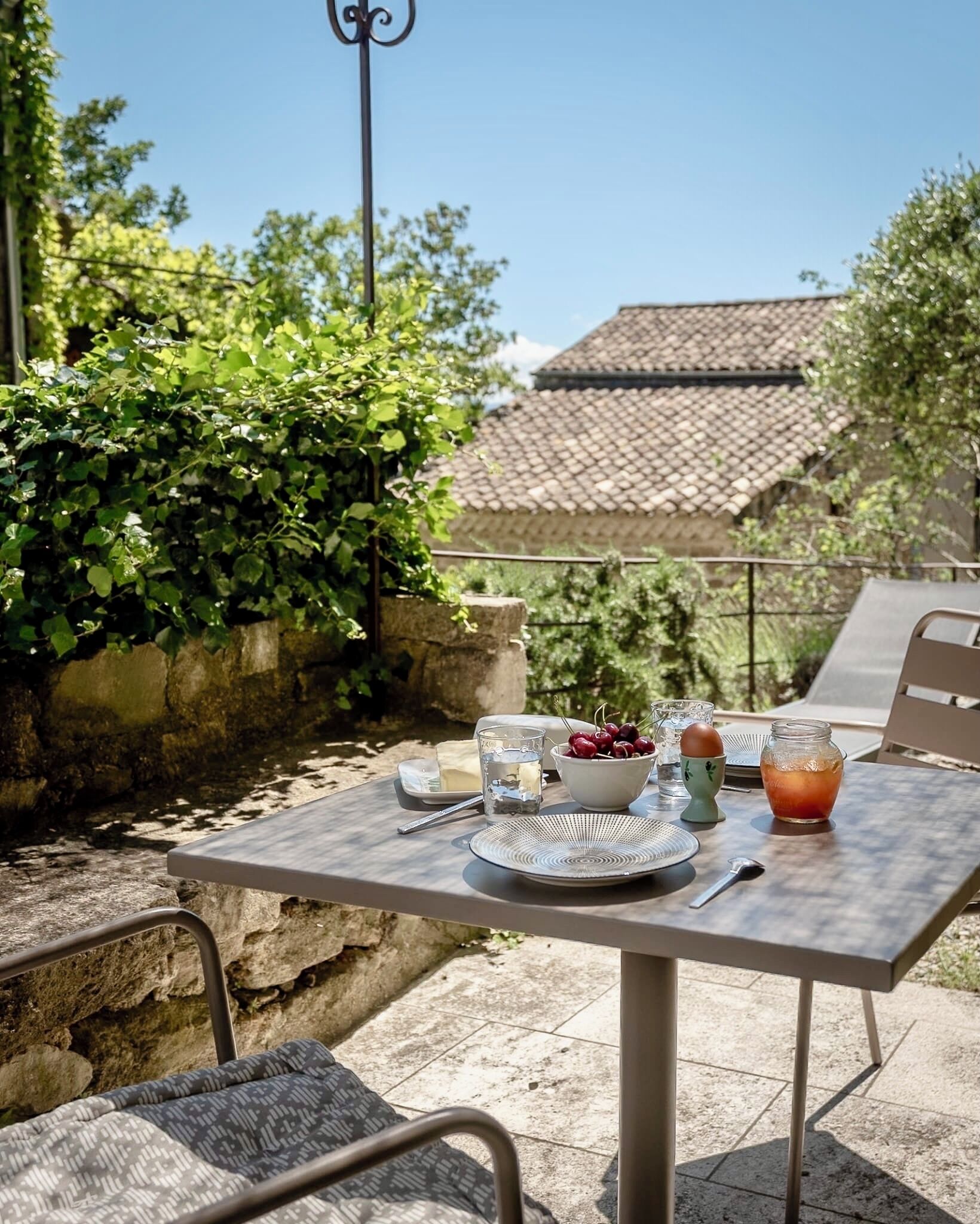 Une table à manger installée à l’extérieur, avec des assiettes, des verres, un bol de cerises et une carafe de thé glacé.