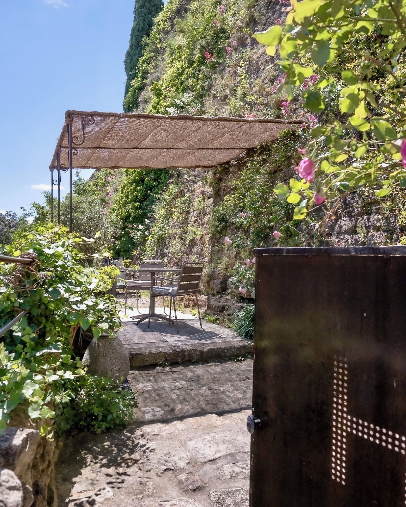 Ein Steinweg führt zu einer Bank unter einer Pergola, umgeben von Grünpflanzen und einer felsigen Klippe.