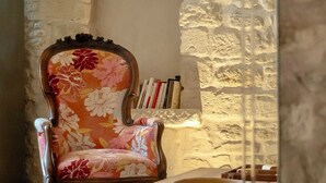 Una habitación con pared de piedra, una silla de madera tapizada con motivos florales, una mesa de madera y estanterías.