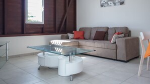 A modern living room with a glass-top coffee table, a beige sofa with cushions, a glass dining table, and a window with curtains. - Villa F3 standing and all comfort in Petit-Bourg in Guadeloupe (PETIT-BOURG)