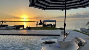 Ett poolområde vid vattnet med solstolar, en möblerad sittgrupp under tak och utsikt över solnedgången över vattnet.