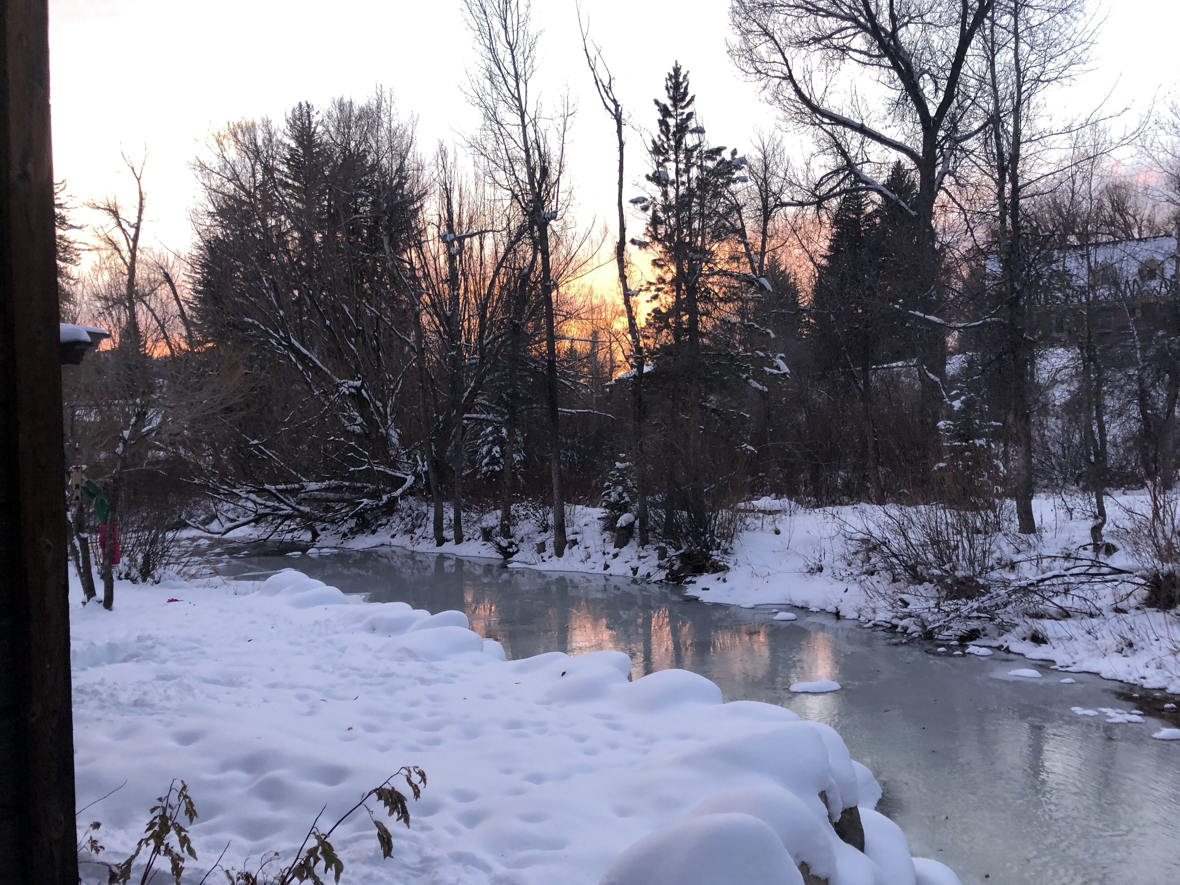 Paisaje nevado con un río congelado, árboles y una puesta de sol al fondo.