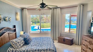 Une chambre avec un grand lit, un ventilateur de plafond, une commode en bois et une vue sur une piscine.