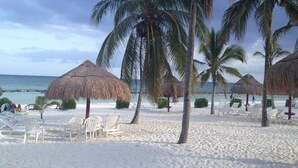 Une plage avec des chaises longues blanches, des palmiers et des parasols en feuilles de palmier.
