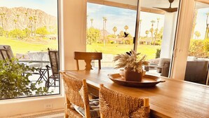 A dining area with a wooden table, woven chairs, and a view of a landscaped garden through large windows. - Breathtaking views from your private pool & spa in PGA West: 3 BD/STR #065869 (La Quinta)