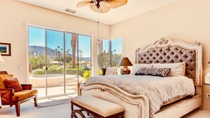 A bedroom with a large bed, a bench, a chair, and a view of palm trees and mountains through large sliding doors. - Breathtaking views from your private pool & spa in PGA West: 3 BD/STR #065869 (La Quinta)
