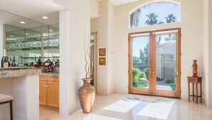 A spacious foyer with a large glass door leading to a garden, a marble floor, and a kitchen area with a glass backsplash and a vase of flowers. - Breathtaking views from your private pool & spa in PGA West: 3 BD/STR #065869 (La Quinta)