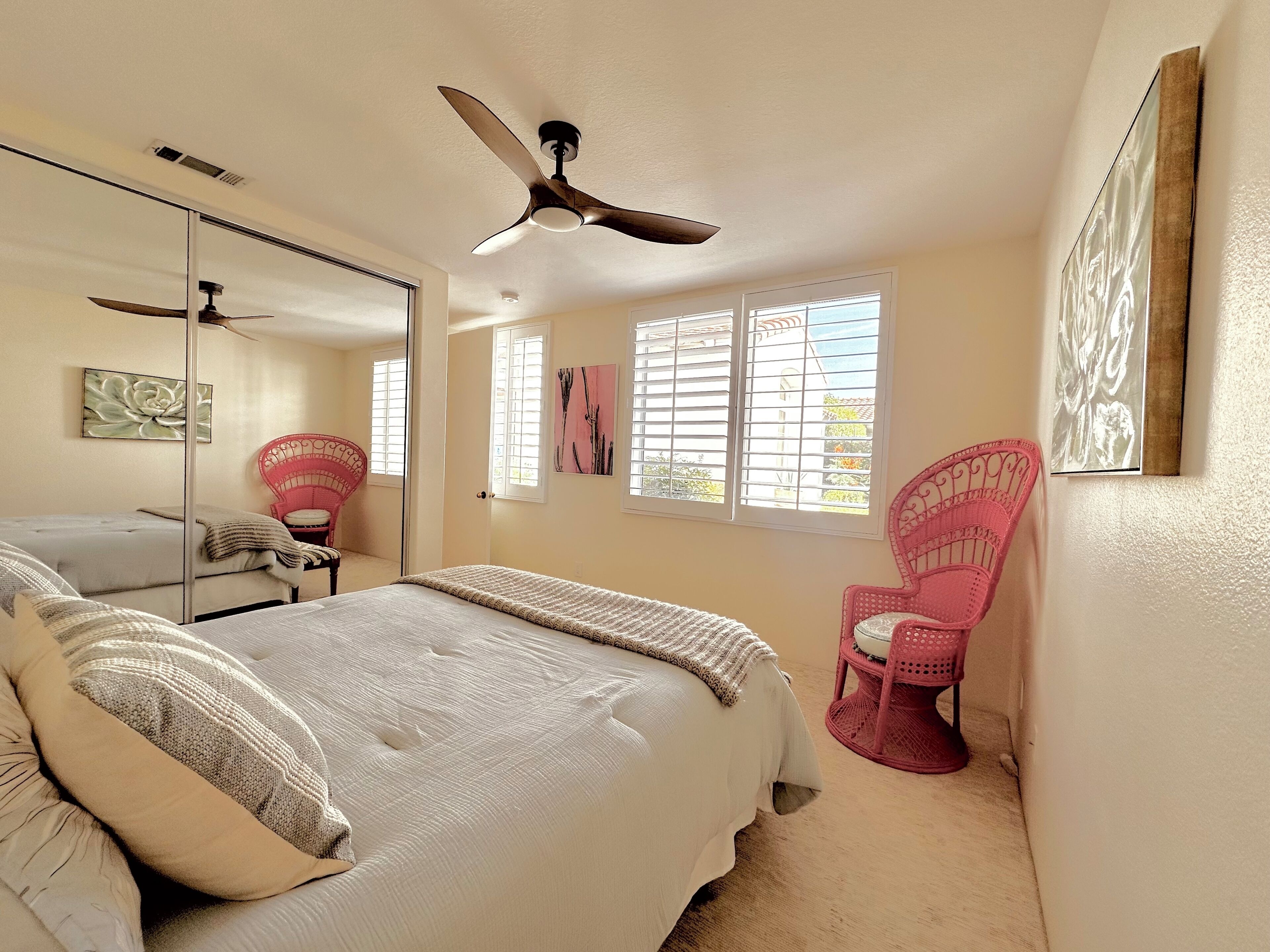 Un dormitorio con cama, ventilador de techo, ventana con persianas, espejo y una silla rosa.