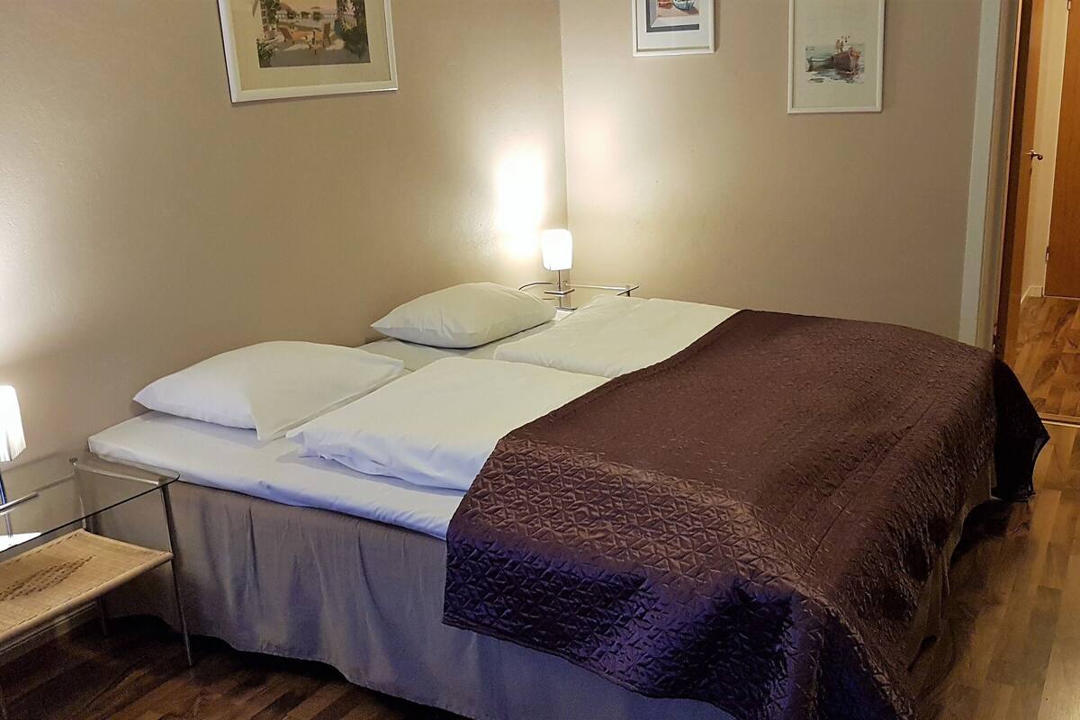 A neatly made bed with white linens and a brown blanket, a bedside table with a lamp, and two framed pictures on the wall.