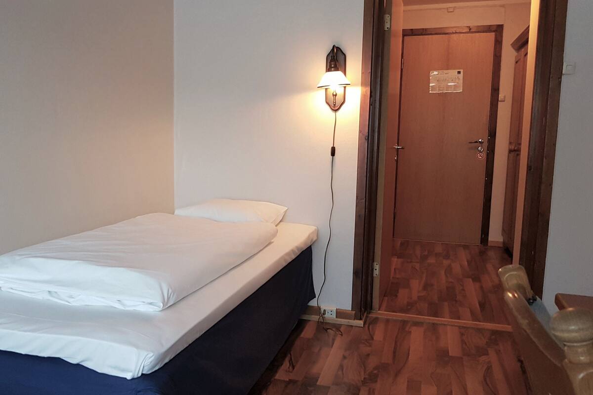A single bed with white linens in a room with wooden flooring and a wall-mounted light fixture.