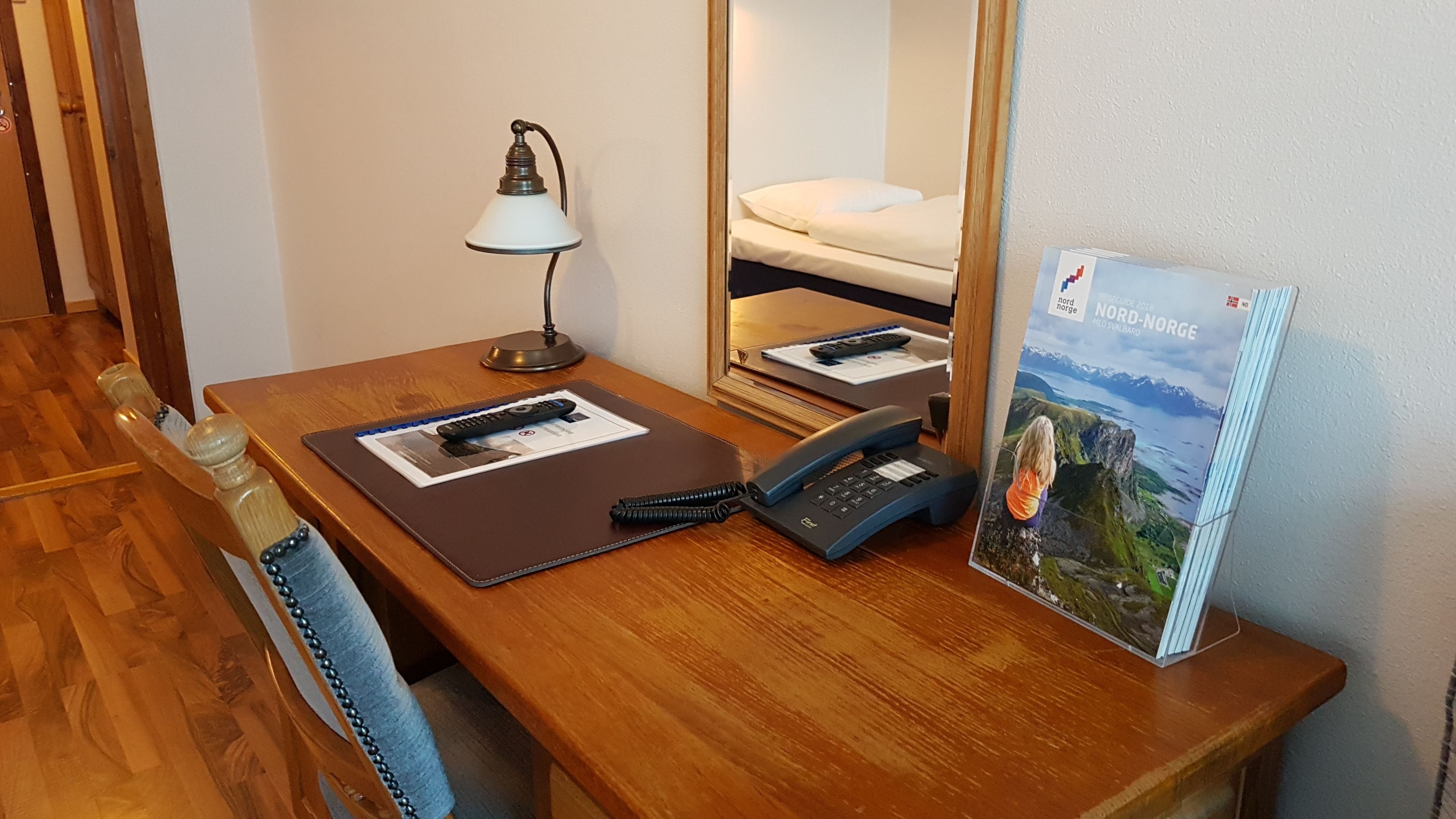 Habitació d'hotel amb escriptori de fusta, un telèfon, un comandament a distància, una revista i un llit amb matalàs i almoades blanques.