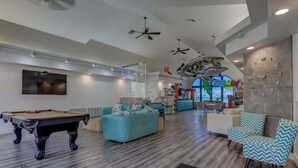 A modern living room with a pool table, a sofa, and a coffee table. There is a kitchen area in the background with a large window. - Las Palmas Resort #1627 | All-Inclusive Amenities (Saint George)