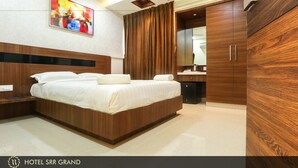 A modern hotel room with a large bed, wooden headboard, a ceiling fan, recessed lighting, a painting on the wall, and a wardrobe. - Hotel SRR Grand (Chengalpattu)