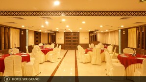 A banquet hall with round tables, white chairs, and red tablecloths. - Hotel SRR Grand (Chengalpattu)