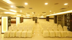 A conference room with rows of white chairs, a stage at the front, and a door leading to another room. - Hotel SRR Grand (Chengalpattu)