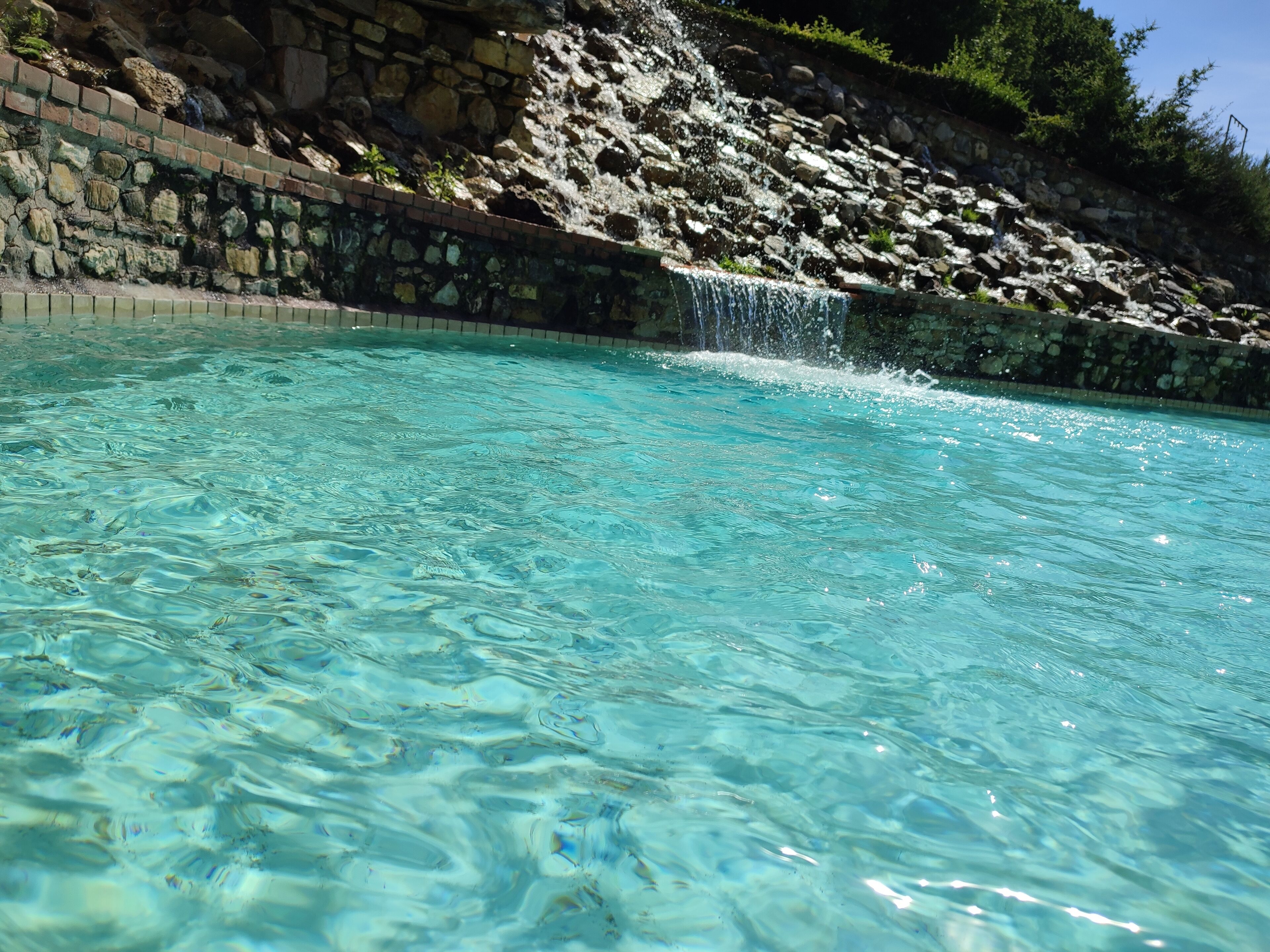 Una piscina cristallina con cascata, circondata da un muro in pietra e vegetazione.