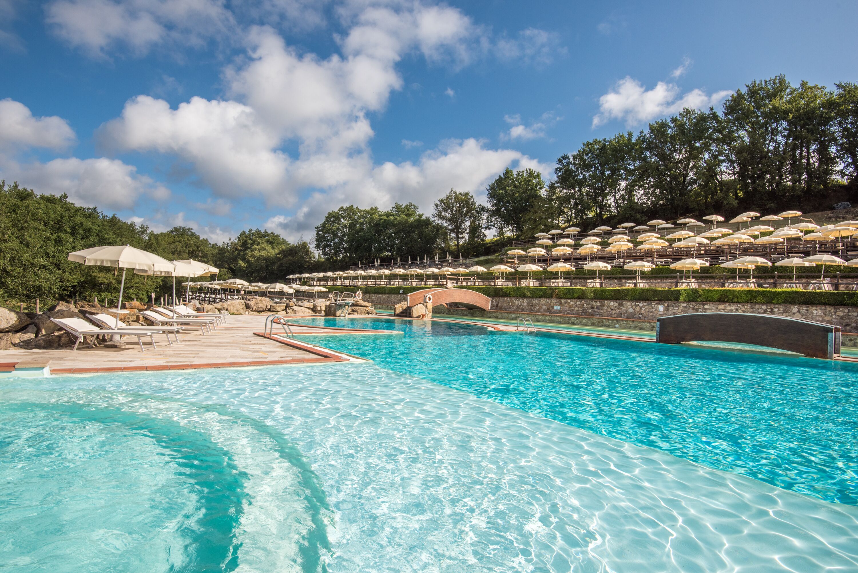 A swimming pool with lounge chairs and umbrellas, surrounded by trees and a stone wall.