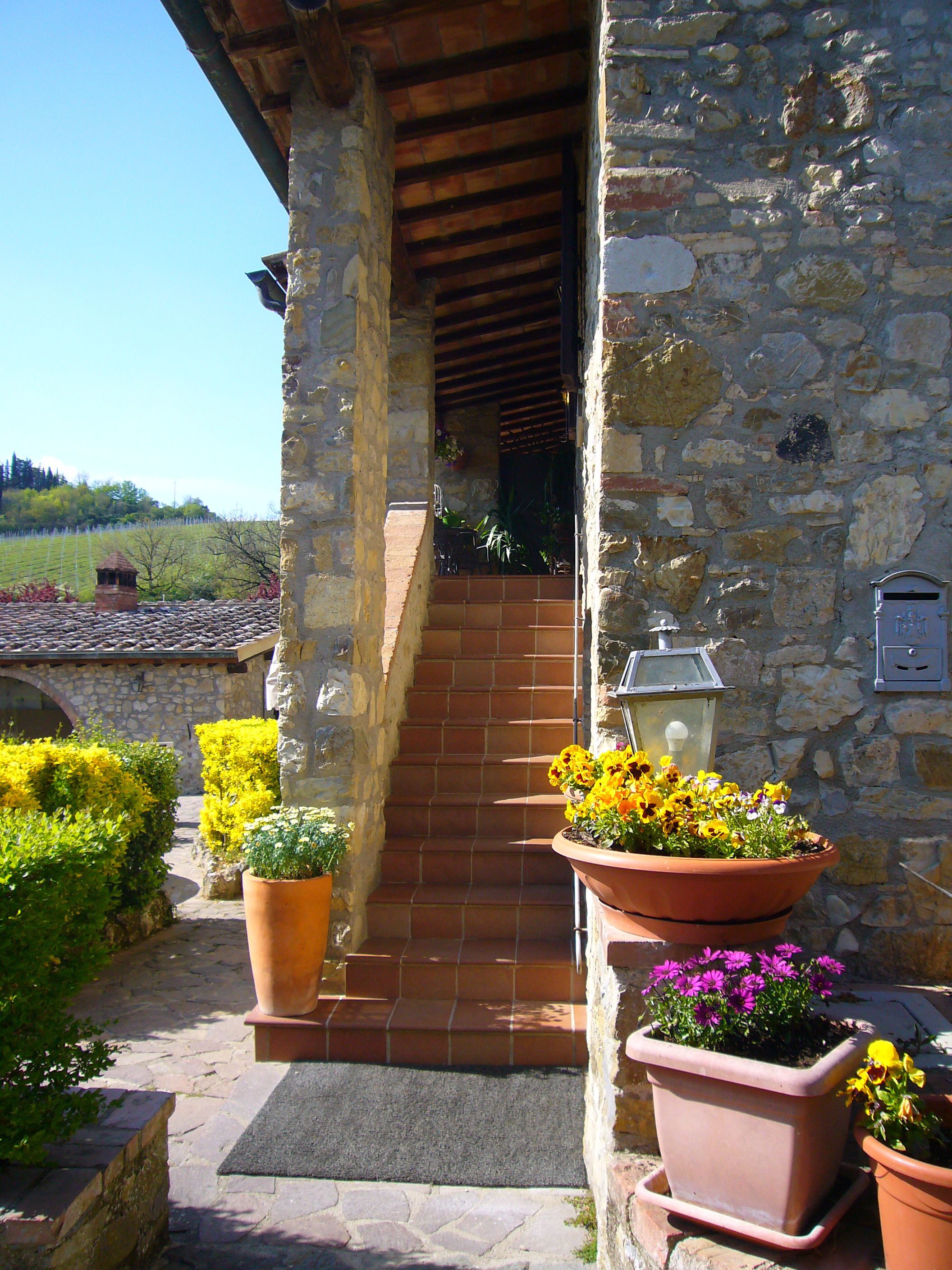 A stone building with a wooden staircase leading to a covered entrance. There are terracotta pots with flowers and a lantern on the steps.