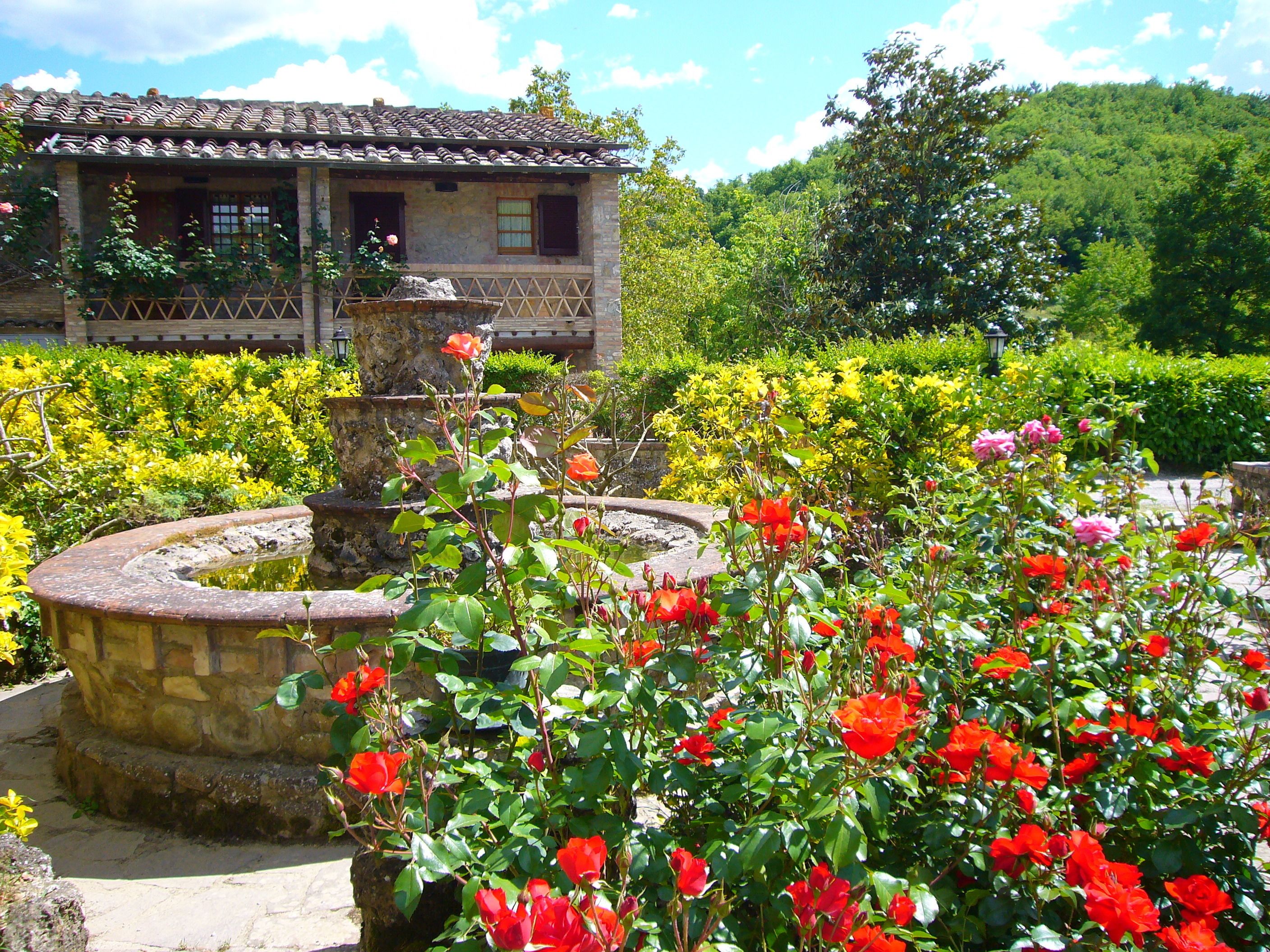 Un edificio de piedra tradicional con techo de tejas, rodeado de un exuberante jardín con rosas rojas vibrantes y flores amarillas.