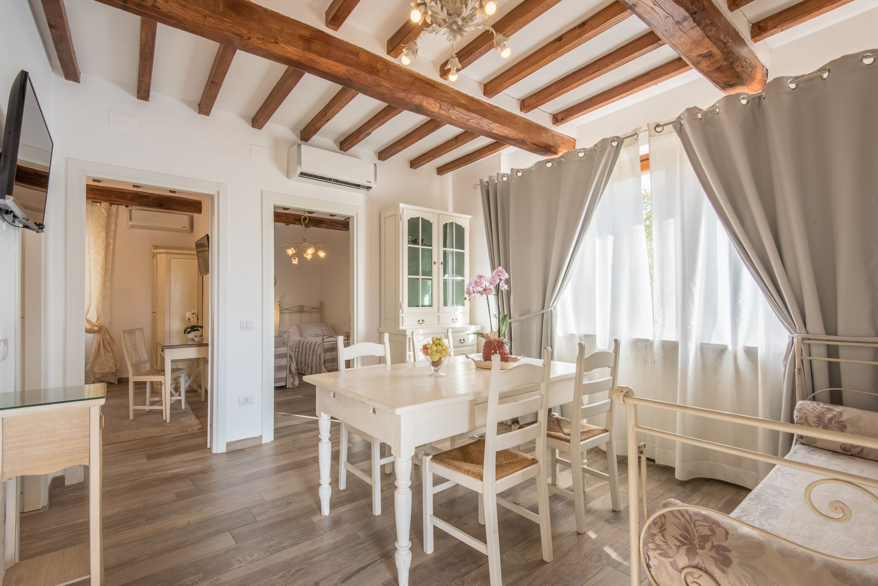 A room with a wooden ceiling, a dining table with chairs, a bed with a headboard, and a window with curtains.