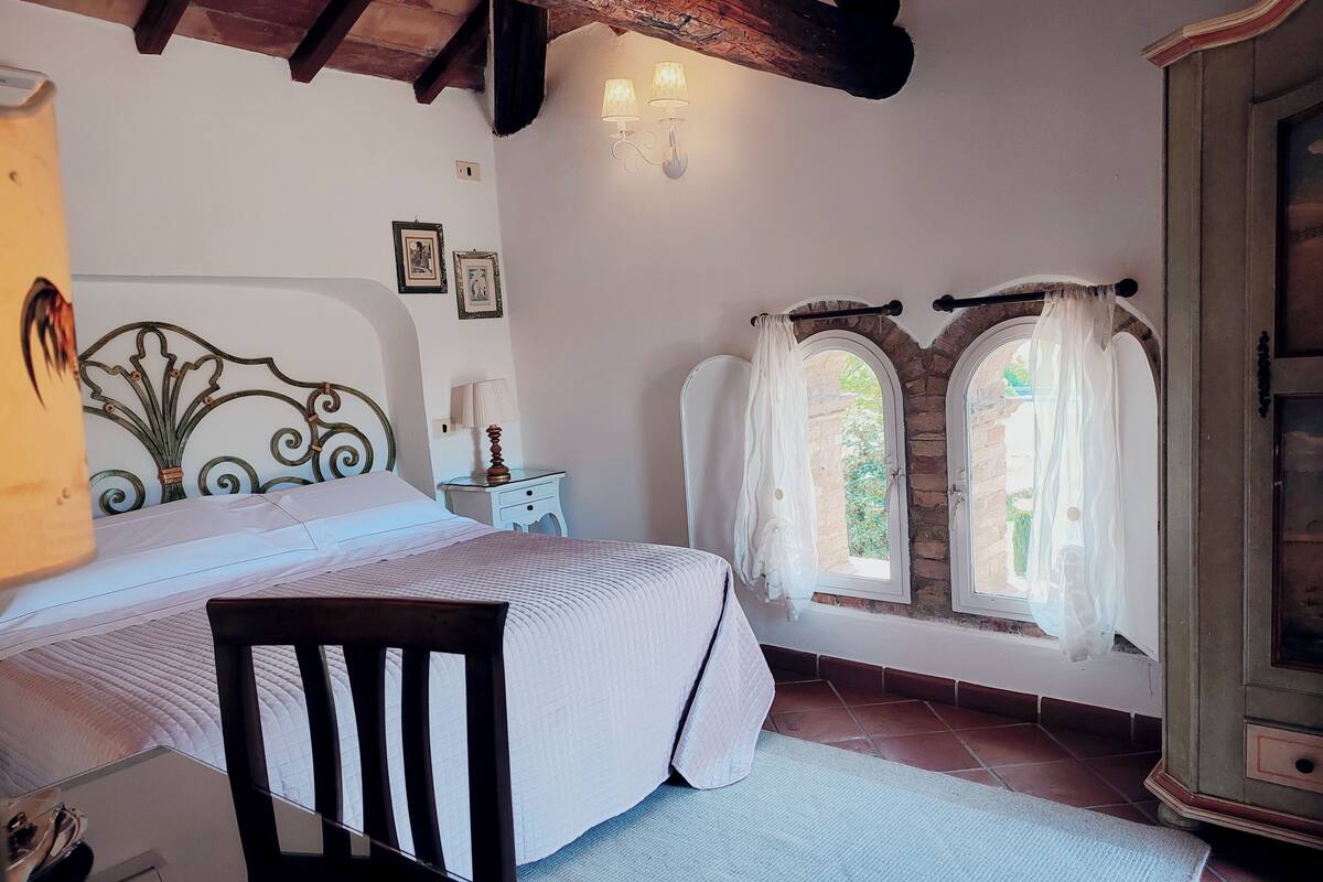 Un dormitorio con cama, una silla, una mesita de noche, una lámpara, un armario y dos ventanas con cortinas.