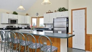 A kitchen with white cabinets, a stainless steel refrigerator, a microwave, and a coffee maker. There are four chairs around a central island. - Walk to the beach, Hot Tub, Rec room, private pool ! (Corolla)