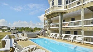 A poolside area with a wooden deck, white lounge chairs, and a multi-level building with balconies. - Walk to the beach, Hot Tub, Rec room, private pool ! (Corolla)
