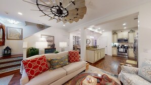 Ein geräumiges Wohnzimmer mit einem beige farbenen Sofa, gemusterten Kissen und einem außergewöhnlichen Deckenventilator. Im Hintergrund sind eine Küche mit modernen Geräten sowie ein Essbereich mit einer Chandeliebre sichtbar.