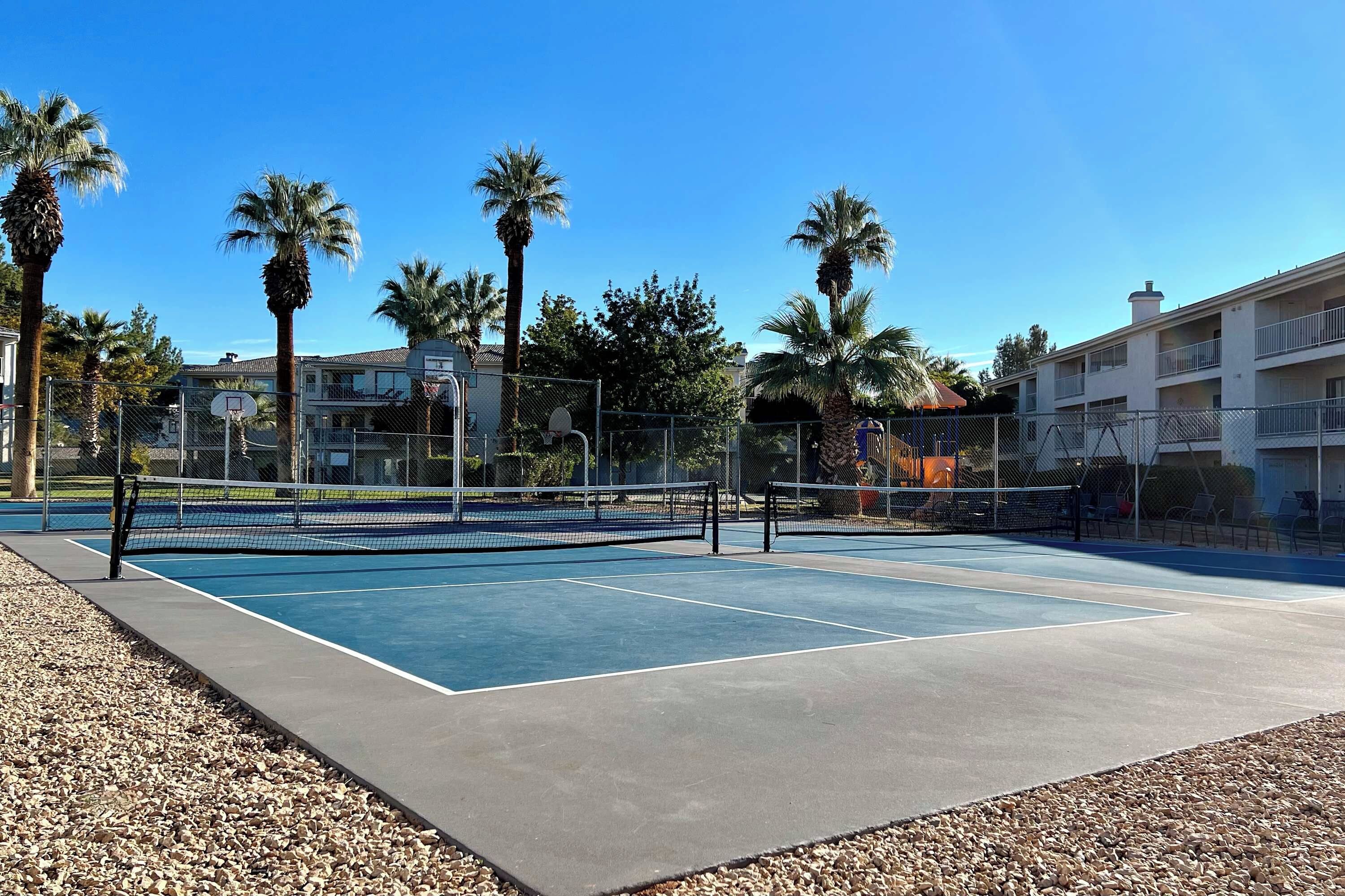 An outdoor tennis court with a net, surrounded by palm trees and a fence.